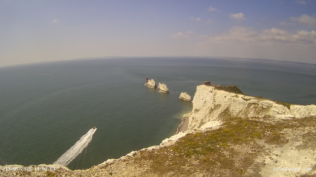 A view of white chalk cliffs with a grassy top standing on the edge of the sea. Several large rock formations are visible offshore, with one red and white lighthouse on one of the rocks. A boat is leaving a wake in the water, heading away from the cliffs. The sky is partly cloudy with scattered white clouds. The image is timestamped "19.05.2023 14:00:03".