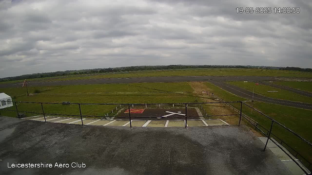 An outdoor view from a webcam at Leicestershire Aero Club shows a cloudy sky over a grassy airfield with runways and taxiways in the distance. The foreground includes a platform with a railing and a small parking area with marked spaces. The time and date displayed on the top right corner are 19-05-2025 at 14:00:50.