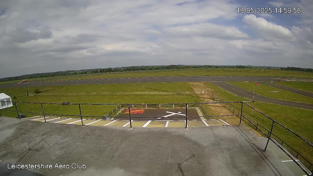 A view from a webcam showing the runway of Leicester Shire Aero Club on a partly cloudy day. The foreground includes a small parking area with marked spaces and a railing, while the background features a wide grassy field, taxiways, and the open sky with scattered clouds. The timestamp indicates the date and time as 19-05-2025 at 14:59:56.