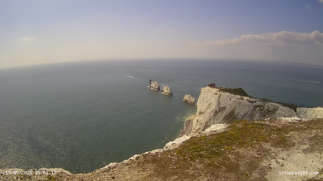 A view from a high vantage point overlooking white chalk cliffs extending into the sea, with a small building or structure on top of the closest cliff. There are rocky formations in the water further out, and a boat or ship is passing by, creating a streak of wake behind it. The sky is mostly clear with a few scattered clouds. The scene is bathed in daylight, and the ocean stretches to the horizon.