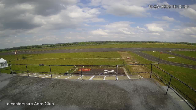 A wide view of an airfield with a grassy area and a runway extending into the distance under a partly cloudy sky. In the foreground, there is a parking area with a white aircraft shelter and a helipad marked with a white "H" on a red background. The image is timestamped "19-05-2025 16:00:00" in the upper right corner, and "Leicestershire Aero Club" is written in the bottom left corner.