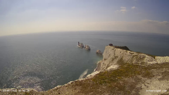 A view of white chalk cliffs rising from the ocean with a small building and a lighthouse on top. Several large sea stacks are visible offshore, and the sea sparkles under the sunlight. The sky is mostly clear with a few scattered clouds. The timestamp in the bottom left corner shows "19/05/2025 16:00:36."