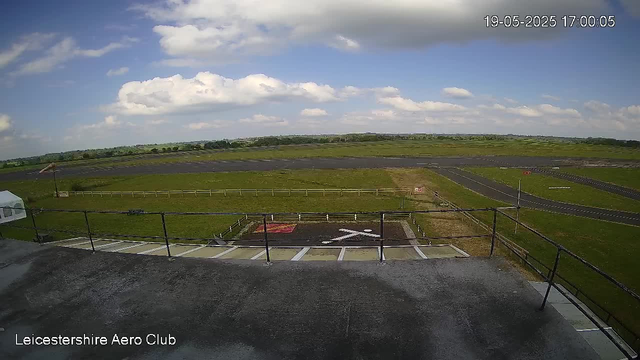 A view from a webcam at LeicesterShire Aero Club showing a clear sky with scattered clouds over a grassy airfield. The foreground includes a small parking area with striped parking bays and a helicopter landing pad marked with a white "X". There are some railway tracks running across the middle distance. The scene appears calm and bright, with the time and date stamp reading 19-05-2025 at 17:00:05.