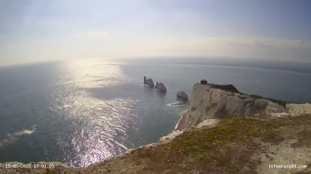 A view from a coastal cliff looking out over the ocean with the sun shining brightly, creating reflections on the water. In the distance, there are large rock formations rising out of the sea, and a small building or lighthouse is situated on top of the cliff. The sky is mostly clear with some light clouds, and the overall scene is serene and sunlit. The timestamp in the lower left corner indicates the date and time as May 19, 2025, at 17:01:05.