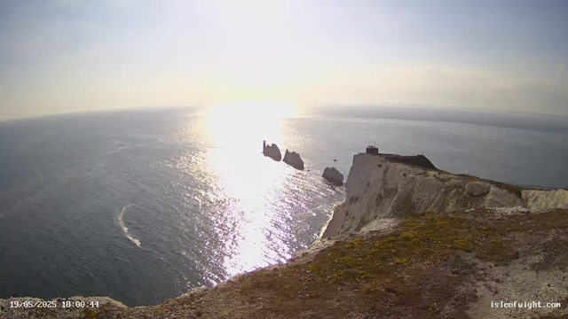 A view of the ocean with white chalk cliffs in the foreground. The sun is setting over the water, reflecting bright light across the surface. There are large rocks jutting out of the sea, and two small buildings or structures can be seen on the cliff's edge. The overall scene is calm and illuminated by the golden glow of the sunset.