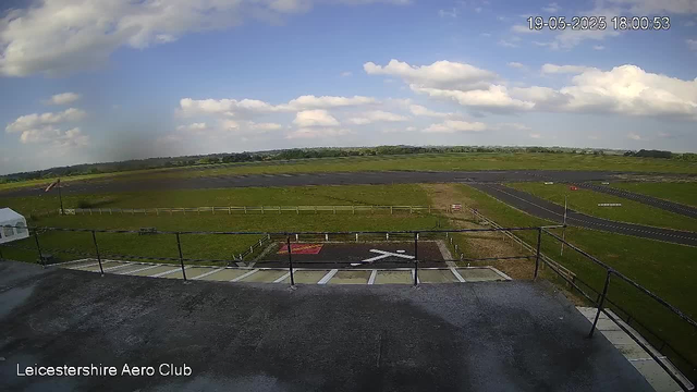 An outdoor view from a webcam at Leicester Shire Aero Club showing a grassy field with a racetrack or runway in the distance under a partly cloudy sky, with a timestamp of 19-05-2025 18:00:53 in the top right corner. The foreground includes a paved area with marked parking spaces and a black railing.