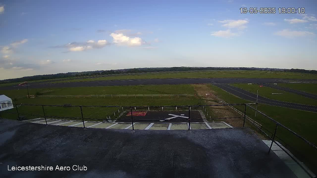 An outdoor view of an airfield on a clear day with blue skies and some scattered clouds. The image shows a grassy area with a runway stretching across the background. In the foreground, there is a helipad marked with a large white "H" and surrounded by a small fenced area, along with some parking spaces. The timestamp on the image reads "19-05-2025 19:00:10," and the bottom left corner notes "Leicestershire Aero Club."