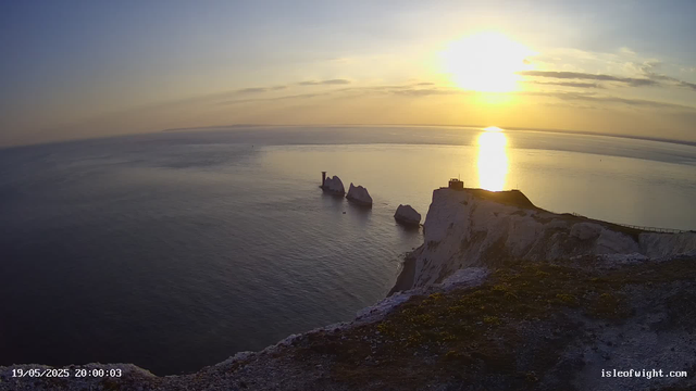 A view of white chalk cliffs overlooking the ocean at sunset. The sky is partly cloudy with the sun low on the horizon, casting a golden reflection on the water. There are large rock formations and a small building on top of the cliffs in the foreground.