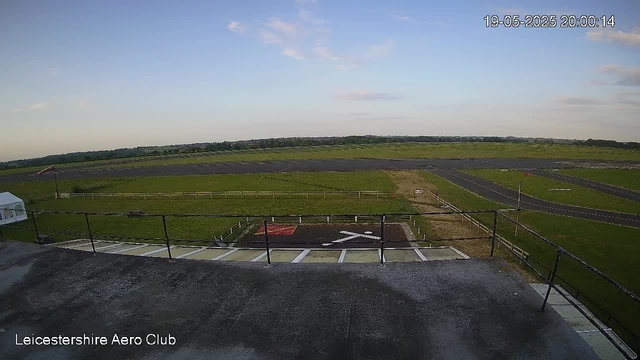 An empty aircraft tarmac at Leicestershire Aero Club under a partly cloudy sky during daytime. The view is from a high vantage point, showing a fenced helipad with a white "H" marking, surrounding grassy areas, and a runway in the distance. The timestamp indicates it was taken on May 19, 2025, at 8:00 PM and 14 seconds.