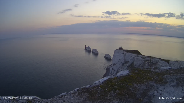 A view of white chalk Cliffs of Dover at sunset, with the sea in the background. Several large sea stacks and rock formations are visible near the coast, with a small watchtower or building perched on the highest point of the cliffs. The sky is partly cloudy with hints of pink and purple as the sun sets. The timestamp indicates the date and time as May 19, 2025, at 21:00:07.
