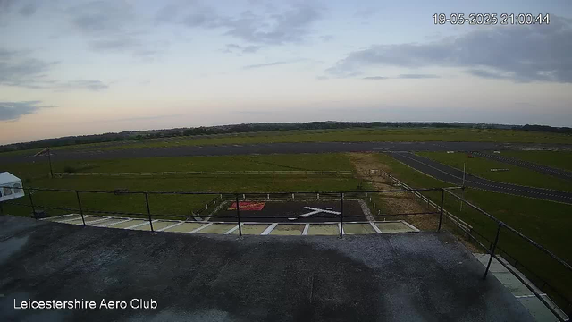 An outdoor view from a webcam showing an airfield with a runway stretching into the distance under a partly cloudy sky. In the foreground, there is a rooftop or balcony with a safety railing, and a helipad marked with a white helicopter symbol. The bottom left corner has the text "Leicestershire Aero Club," and the top right corner displays a timestamp: "19-05-2025 21:00:44."