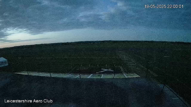 A nighttime view from a webcam at Leicestershire Aero Club. The sky is dark with some clouds, and a faint hint of light is visible on the horizon. The foreground shows the roof of a building and a small helipad area with a helicopter parked on it. The image is slightly dark and grainy. The date and time stamp in the top right corner reads 19-05-2025, 22:00:01.