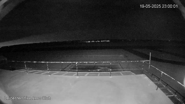 A black-and-white image from a webcam showing an empty outdoor area at night. There is a metal railing or fence in the foreground, with a flat surface behind it. The sky is dark, and a faint line of distant lights is visible on the horizon. The date and time stamp in the upper right corner reads "19-05-2025 23:00:01," and the bottom left corner contains the text "Leicestershire Aero Club."