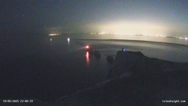 A nighttime scene of a body of water seen from a boat or dock. The horizon is faintly illuminated by distant city lights or lightning, creating a glowing line in the background. There are small, bright reflections on the water's surface, including a prominent red light toward the center-left. The scene is dark with very limited visibility of the surrounding landscape or objects. The image has a timestamp in the bottom left corner and a website URL in the bottom right corner.