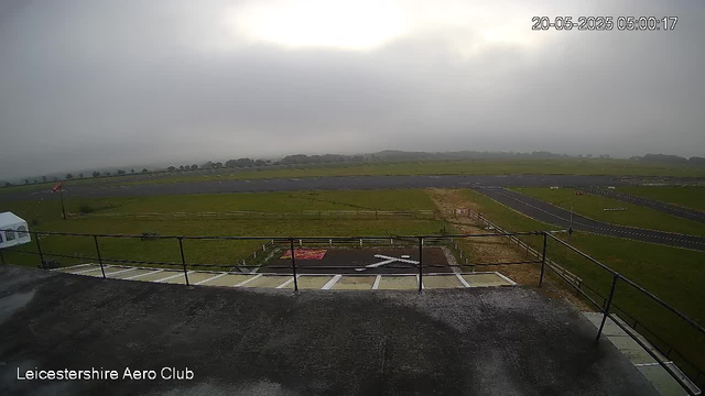 A view from a webcam overlooking an airfield on a cloudy day. The foreground shows a concrete balcony with a black railing, a helicopter landing pad marked with a white "H," and some parking spaces. Beyond the balcony, there is a grassy area, taxiways, and runways stretching into the distance. The sky is overcast with dark clouds, and the landscape appears misty. The timestamp on the image indicates it was taken on May 20, 2025, at 5:00:17 AM. The bottom left corner contains the text "Leicestershire Aero Club."