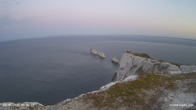 A view of white chalk cliffs overlooking the ocean at dawn. There are three prominent rock formations in the water near the cliffs, including a red and white lighthouse on one of the rocks. A small building is situated on top of the cliffs, with a grassy area surrounding it. The sky is soft pastel colors, and the water appears calm. The image has a timestamp in the lower-left corner indicating early morning, and a website address "isleofwight.com" in the lower-right corner.