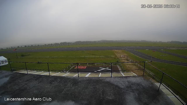 A view from a webcam overlooking an empty aircraft takeoff or landing area on a cloudy day. The scene includes a small section of tarmac, fencing, grassy fields, and a runway stretching into the distance. The image is timestamped "20-05-2025 06:00:48" and labeled "Leicestershire Aero Club."