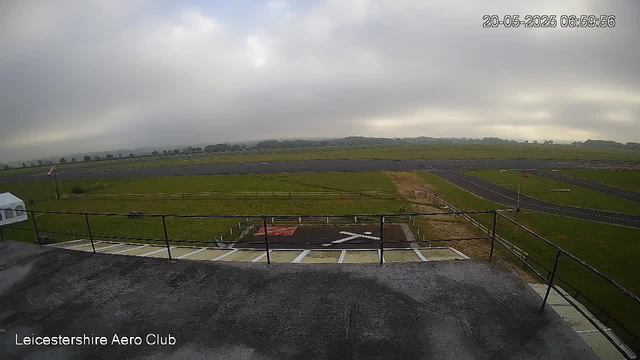 A view from a webcam overlooking an airfield at Leicestershire Aero Club on a cloudy day. In the foreground, there is a parking area with designated spots, some marked with white lines and a large white arrow. Beyond the parking, a grassy area is visible with a small fence and a windsock on a pole. The airfield runway stretches across the middle ground, surrounded by green fields that extend toward a cloudy, overcast sky in the background. The timestamp in the upper right corner indicates the date and time as May 20, 2025, at 6:59:56.