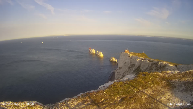 A panoramic view of white chalk cliffs overlooking the ocean, with a rocky outcrop and a lighthouse on the far right. Several sailboats are visible in the water, and the sky is mostly clear with a few wispy clouds. The scene is bathed in warm, golden sunlight.