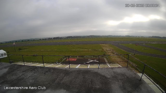 A wide view of an overcast sky over an airport runway and grassy field. The foreground shows a parking area with designated spots and a small helipad marked with a white 'H'. There is a railing along the edge of the parking area. In the top right corner, there is a timestamp reading "20-05-2025 08:00:24". The bottom left corner has text that says "Leicestershire Aero Club".
