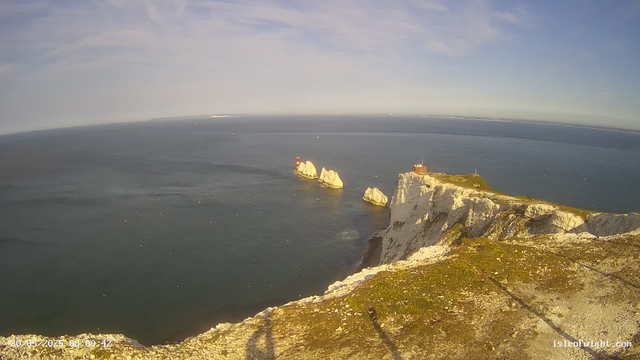 Aerial view of white chalk cliffs with patches of green grass on top, overlooking the ocean. There are several large rock formations protruding from the water near the cliffs. The sky is partly cloudy with sunlight illuminating the scene. A lighthouse or navigation tower is visible on the edge of the cliffs. The photo timestamp shows the date as May 20, 2025, at 8:00:42 AM.