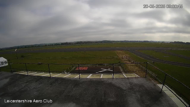 A wide view of an empty airport runway and grassy fields under a cloudy sky, captured from a webcam at Leicesterhire Aero Club. The image shows a fenced platform, taxiways, and a large open airfield extending into the distance. The timestamp in the top right corner reads "20-05-2025 09:00:45."