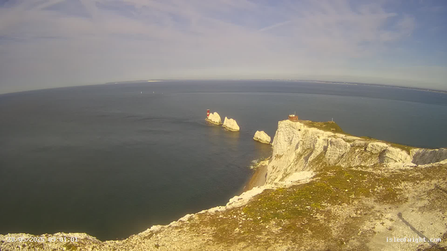 A wide view of a coastal landscape showing white chalk cliffs extending into the ocean, with several large rock formations emerging from the water near the cliffs. A small building and a lighthouse are situated on top of the cliffs, with a clear blue sky and some wispy clouds overhead. In the distance, sailboats are visible on the water.