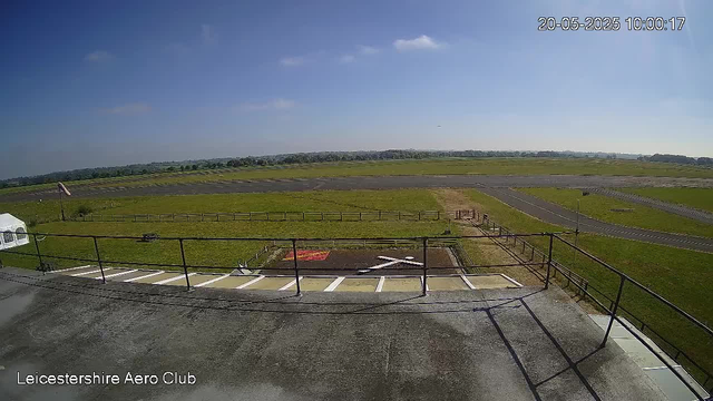 An outdoor view from a webcam showing a flat, grassy airfield with a runway extending into the distance. There is a small parking area with marked parking spaces and a white tent on the left side. A flagpole with a flag is visible near the tent. The sky is mostly clear with a few scattered clouds, and the weather appears sunny. The time and date stamp in the top right corner reads "20-05-2025 10:00:17," and the bottom left corner displays "Leicestershire Aero Club."