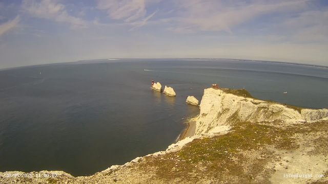 A panoramic view of white chalk cliffs along the coastline under a partly cloudy sky. The cliffs extend into the sea, with a small red and white lighthouse situated on one of the rocks. Several boats are visible in the water, and the horizon shows a distant landmass. The foreground includes the top of the cliff covered in patches of moss and dirt. The image has a timestamp and website URL at the bottom.