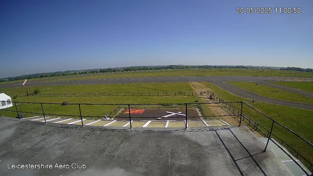 An empty runway with a grassy field and a clear blue sky in the background. A small helipad with white markings is visible in the foreground. A black metal railing runs along the edge of the viewing platform, with parking spaces and a small trailer to the left. The date and time stamp in the top right corner reads "20-05-2025 11:00:50." Text in the bottom left corner reads "Leicestershire Aero Club."