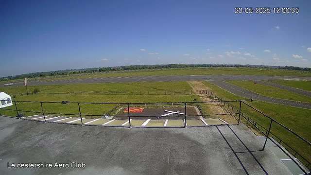 A panoramic view of an airfield under a clear blue sky with a few scattered clouds. In the foreground, there is a helipad with a white "H" marked on it, surrounded by a small fence and a red-green wind sock nearby. The airfield has grassy areas and paved runways extending into the distance. A fence borders the grassy areas, and the horizon shows a mix of fields and trees. The timestamp in the top right corner reads "20-05-2025 12:00:35." The bottom left corner displays the text "Leicestershire Aero Club."