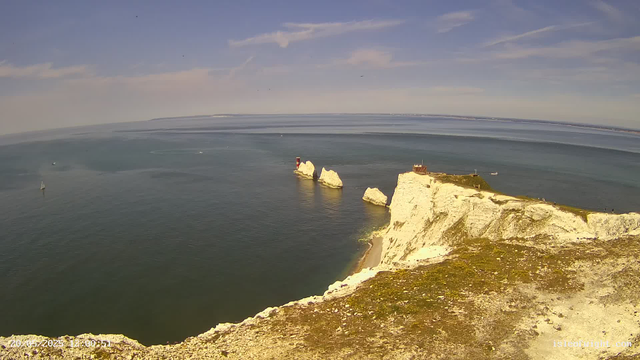 Aerial view of white chalk cliffs with a grassy top, jutting into the sea. There is a small building and a lighthouse on the cliff's edge. The sea below is calm with a sailboat and a few other boats visible. The sky is partly cloudy with patches of blue.