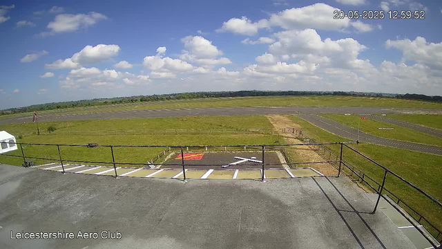 A wide view of an airfield under a bright blue sky with scattered white clouds. The image shows a runway stretching into the distance, surrounded by green grass. In the foreground, there is a small parking area with white lines, and an orange and black marked helipad with a white helicopter symbol. The date and time are displayed in the top right corner as "20-05-2025 12:59:52". The bottom left corner has the text "Leicestershire Aero Club".