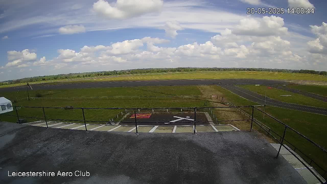 A wide view from a webcam showing a grassy airfield with runways and taxiways under a partly cloudy sky. In the foreground, there is a balcony with a black railing, and a helicopter landing pad marked with a white "H" and an arrow. The time and date are displayed in the upper right corner as "20-05-2025 14:00:44." The bottom left corner has the text "Leicestershire Aero Club."