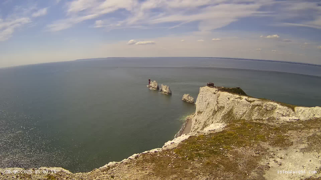 A view from a webcam showing white chalk cliffs extending into the sea with rock formations emerging from the water. The sky is partly cloudy with streaks of clouds across a blue sky, and the ocean appears calm with gentle waves. The foreground has a rocky and grassy edge of the cliff. The timestamp in the lower left corner indicates the date and time as May 20, 2025, at 15:00:15.