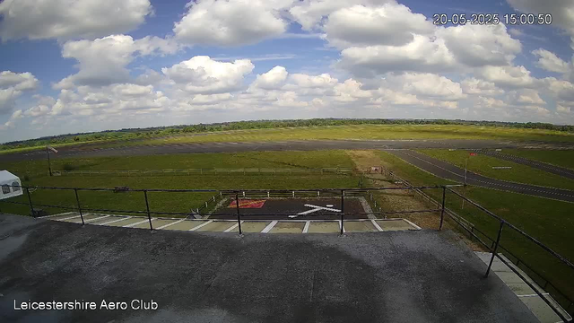 A wide view of an airfield during daytime, with a large open grassy area, a runway in the distance, and a partly cloudy sky overhead. In the foreground, there is a landing pad with a white arrow and a red square, surrounded by a black railing. The timestamp in the top right corner reads "20-05-2025 15:00:50," and the bottom left corner displays the text "Leicestershire Aero Club."
