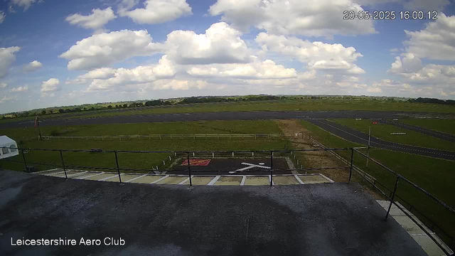 A view from a webcam at Leicester Aero Club showing a partly cloudy sky with large white clouds and patches of blue. In the foreground, there is a rooftop with a railing, a helicopter to the left, and designated parking positions below. The scene extends to open green fields and a runway in the distance, with a few small buildings or structures. The timestamp on the image reads "20-05-2025 16:00:12."