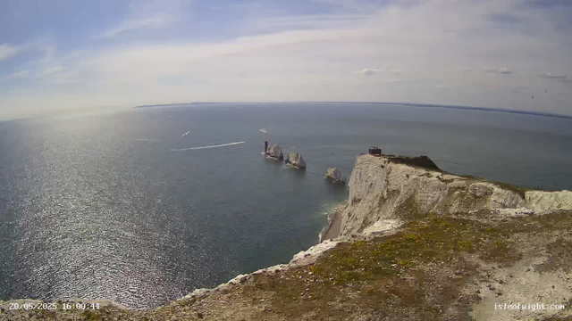 A high-angle view of white chalk cliffs overlooking the ocean, with several rock formations rising from the water near the base of the cliffs. The sea is shimmering under the sunlight, and a few boats are visible in the distance. The sky is partly cloudy with streaks of blue. A small building or structure is situated on top of the cliff. The timestamp at the bottom left reads "20/05/2025 16:00:44."