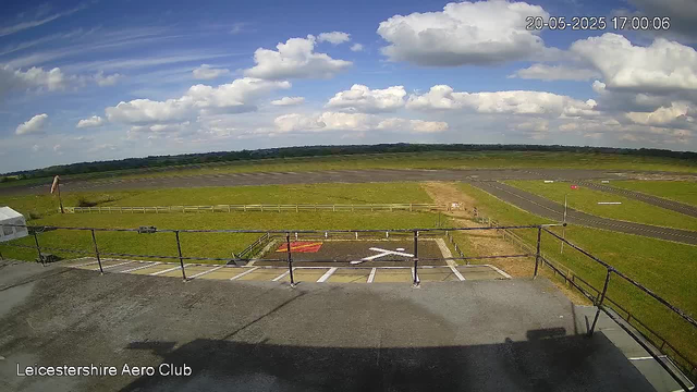 A wide view of an open grassy airfield under a partly cloudy sky, with a small helipad marked with an arrow and landing pad in the foreground, and a stretch of runway extending into the distance. The image is labeled "Leicestershire Aero Club" at the bottom left corner, and the timestamp in the top right corner reads "20-05-2025 17:00:06."