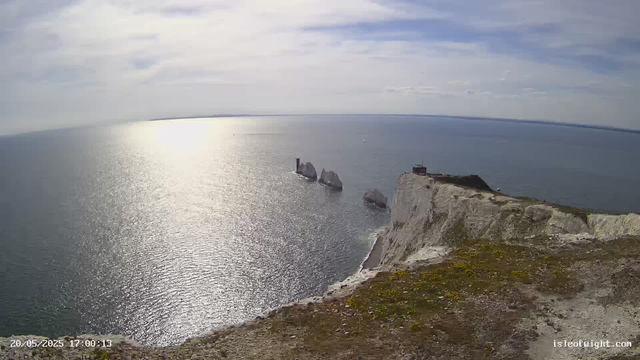A high-angle view of white chalk cliffs extending into the sea, with a small structure on the edge. The ocean is calm with sunlight reflecting off the surface, and the sky overhead has scattered clouds. In the distance, there are some large rock formations rising from the water near the cliffs. The image has a timestamp in the bottom left corner and the website name "isleofwight.com" in the bottom right corner.