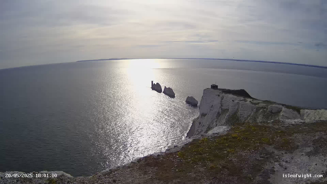 An aerial view of a coastal scene showing white chalk cliffs with a small building and a lighthouse on top, overlooking the ocean. Three large sea stacks rise from the water near the cliffs, with sunlight reflecting off the calm surface of the sea. The sky is partly cloudy. The timestamp on the image reads "20/05/2025 18:01:00" and the website "isleofwight.com" is visible in the bottom right corner.