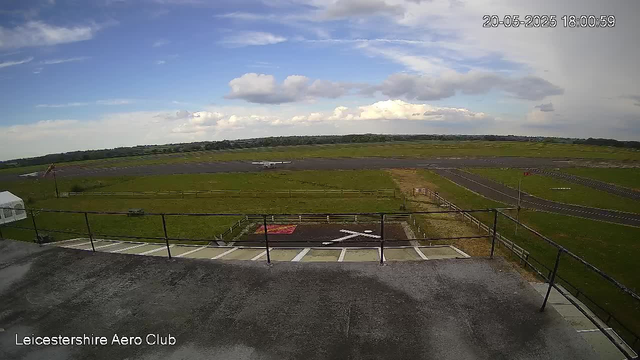A view of an airfield taken from a webcam. In the foreground, there is a concrete terrace with a black metal railing, and below it are marked parking spaces. Beyond the terrace, a grassy area and a runway are visible, with a small aircraft in the distance. The sky is partly cloudy with patches of blue, and the photo is timestamped May 20, 2025, at 6:00 PM. The location is Leicestershire Aero Club.