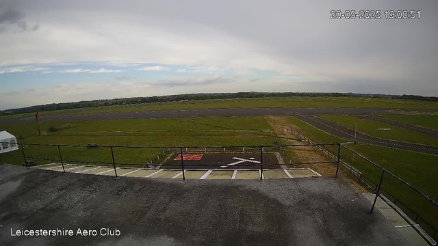 An outdoor view from a webcam at Leicestershire Aero Club showing an empty tarmac runway and taxiway with grassy areas on either side. The sky is partly cloudy with some blue visible, and a distant tree line is seen on the horizon. The image is timestamped "20-05-2025 19:00:51" in the top right corner.