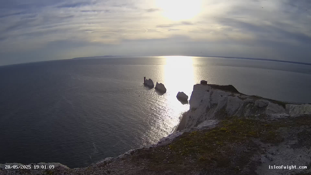 A view of the ocean and white chalk cliffs during sunset. Several large rock formations are visible emerging from the water, with the sunlight reflecting off the surface. The cliff's edge is in the foreground, with patches of green moss or grass. A small structure or building is visible at the top of the cliff on the right side. The image has a timestamp of 20/05/2025 19:01:09 and the website isleofwight.com is displayed at the bottom right corner.