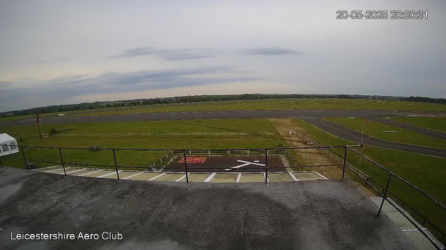 An exterior view of an airfield taken from a webcam. In the foreground, there is a railing and a small parking area with white lines and a red symbol. Beyond that, there are grassy areas and a paved taxiway and runway. The sky is cloudy, and distant trees and buildings are visible on the horizon. The timestamp shows the photo was taken on May 20, 2025, at 8:00 PM. The bottom left corner contains the text "Leicestershire Aero Club."