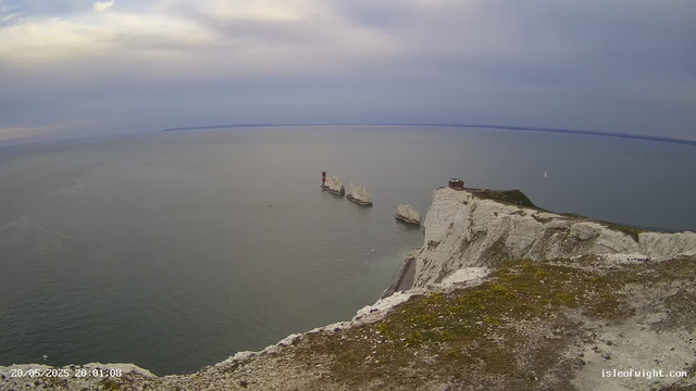A view of white chalk cliffs overlooking the sea, with a small building and a lighthouse near the edge. Several rock formations are visible in the water, and a sailboat is sailing in the distance. The sky is cloudy, and the water appears calm.