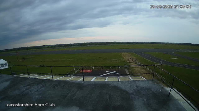 A view from a webcam showing an airfield under a cloudy sky. In the foreground, there is a helipad with a white helicopter symbol and a red marking, surrounded by a metal railing. To the left, part of a glass-walled structure is visible. The middle ground features grassy areas and runway surfaces extending towards the horizon. The timestamp in the top right corner reads "20-05-2025 21:00:06," and the bottom left corner displays the text "Leicestershire Aero Club."