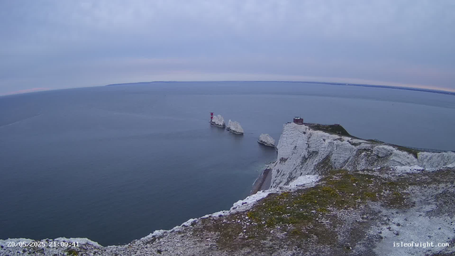 A view of white chalk cliffs extending into the sea, with three large white chalk stacks rising separate from the main cliffs. A small building and a lighthouse are perched atop the cliffs. The sea is calm with a hazy horizon, and the sky is overcast with clouds. The date and time stamp in the bottom left corner reads "20/05/2025 21:00:41," and the bottom right corner shows the website "isleofwight.com."