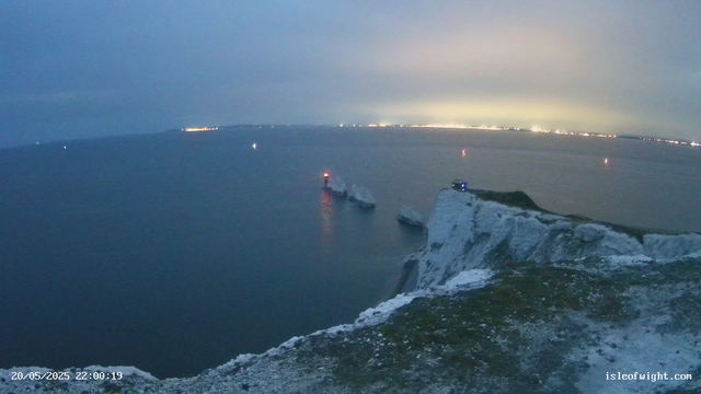 A nighttime view of a coastal cliff with white chalk formations. There are three lighthouse beacons on the cliff emitting red light. The sea below is calm, with a ship anchored near the shore. In the distance across the water, there is a faint glow of city lights. The timestamp in the lower left corner reads "20/05/2025 22:00:19," and a website URL "isleofwight.com" is visible in the lower right corner.