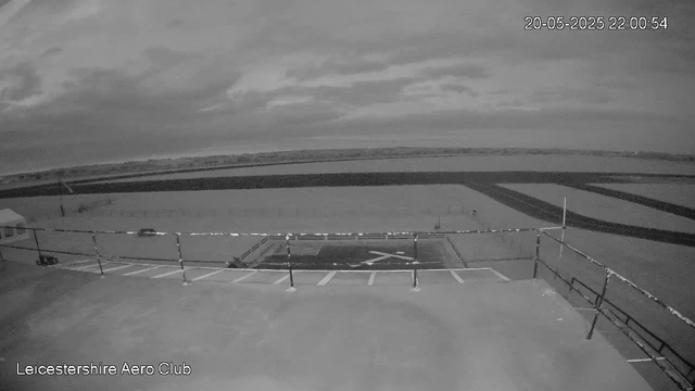 A black-and-white webcam image showing the Leicesterhire Aero Club with a helipad marked by a large "H" at the center. In the foreground, there is an empty parking area with a few parked cars, surrounded by a safety railing. In the background, open fields and a runway or taxiway are visible under a partly cloudy sky. The timestamp in the top right corner indicates the date as May 20, 2025, at 10:00 PM and 54 seconds.